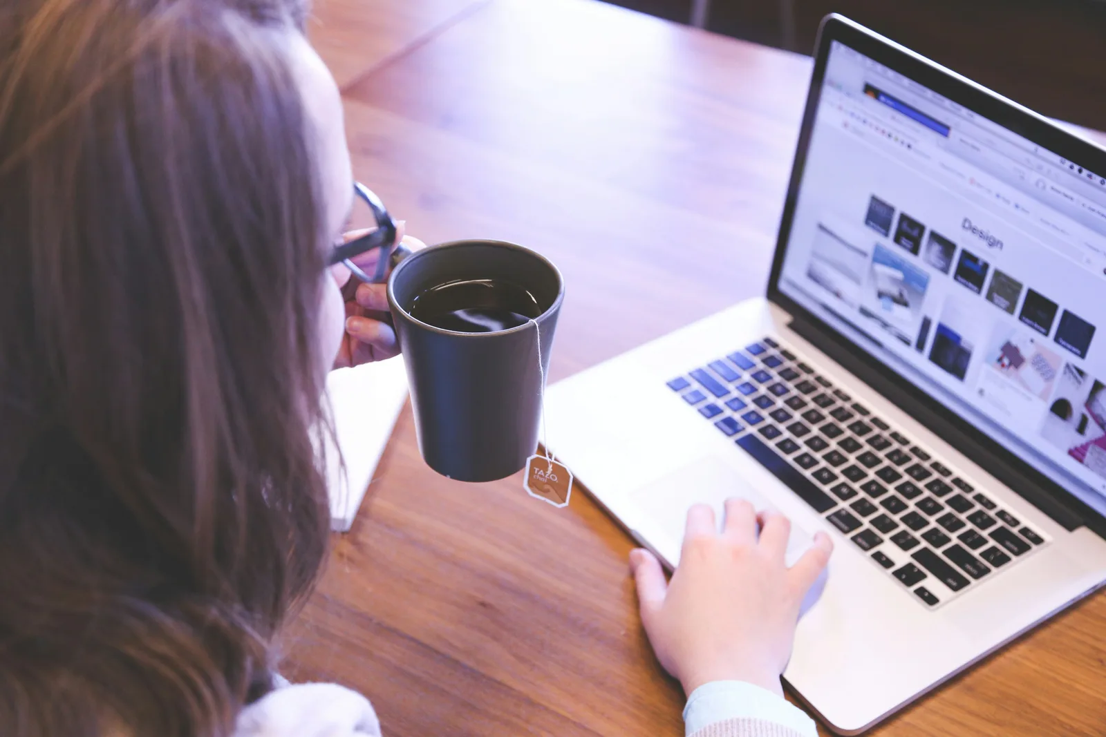 A person with long brown hair, seen from behind, works on a laptop displaying design thumbnails while sipping tea from a black mug with a TAZO tea tag, symbolizing focused content creation for AI SEO.