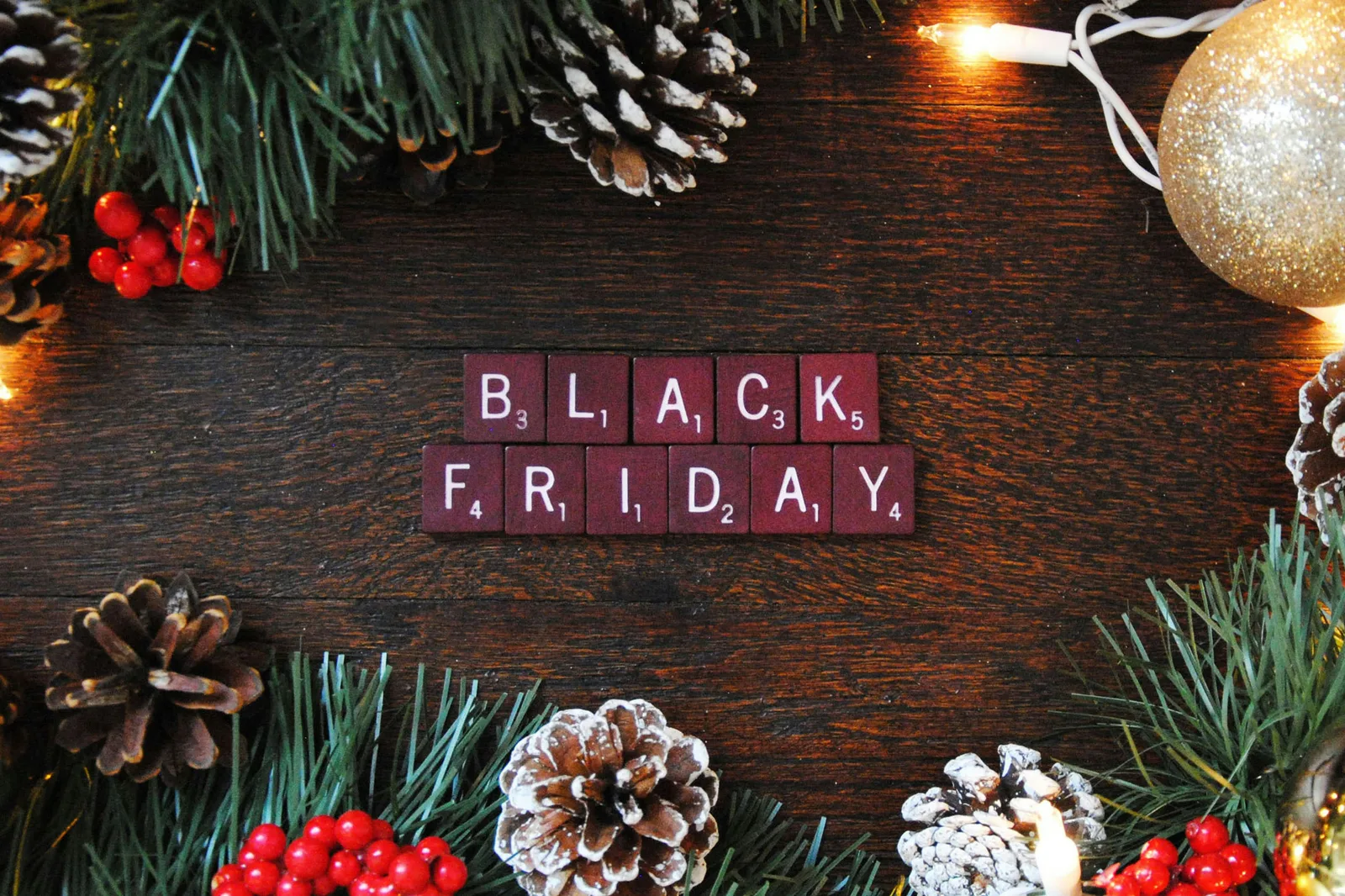 Black Friday spelled out with Scrabble tiles surrounded by pinecones, Christmas lights, and holiday decorations; representing festive season shopping and marketing preparation.